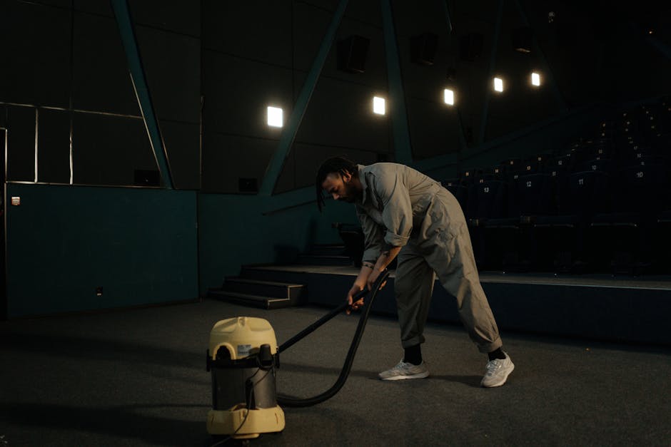 A professional cleaner with dark hair and a beard is operating a yellow vacuum cleaner on a carpeted floor inside a dimly lit auditorium or theater. The cleaner is wearing a light gray jumpsuit and white sneakers, bent over slightly as they vacuum the dark gray carpeted surface. The auditorium features dark blue seating rows with numbered seats and a stage with stairs leading up to it. Overhead, modern square lights are illuminating the seating area, creating a contrast between the bright activity of cleaning and the surrounding darkness. The scene depicts thorough surface cleaning and deep cleaning activities, emphasizing hygienic maintenance within a large, enclosed space and showcasing the professional services offered by Carpet Cleaning Kennington as referenced in the page [PAGE_TITLE].