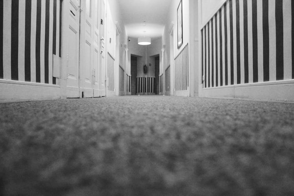 Black and white photograph of a narrow corridor in a residential building, with striped wallpaper on the walls and multiple closed white doors on either side. The carpeted floor appears clean, showing a uniform texture, and the corridor is illuminated by a ceiling light fixture at the far end. The perspective is from a low angle close to the ground, highlighting the surface cleaning and maintenance of the corridor flooring. This image exemplifies the importance of regular deep cleaning and sanitisation of hallways in residential premises, as offered by Carpet Cleaning Kennington, especially important for addressing access issues in basement areas like those described in the SE11, Kennington location.