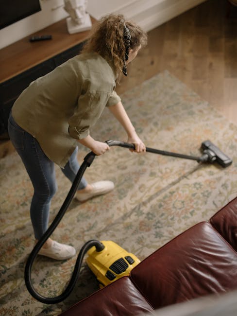 A professional cleaner with dark hair and a beard is operating a yellow vacuum cleaner on a carpeted floor inside a dimly lit auditorium or theater. The cleaner is wearing a light gray jumpsuit and white sneakers, bent over slightly as they vacuum the dark gray carpeted surface. The auditorium features dark blue seating rows with numbered seats and a stage with stairs leading up to it. Overhead, modern square lights are illuminating the seating area, creating a contrast between the bright activity of cleaning and the surrounding darkness. The scene depicts thorough surface cleaning and deep cleaning activities, emphasizing hygienic maintenance within a large, enclosed space and showcasing the professional services offered by Carpet Cleaning Kennington as referenced in the page [PAGE_TITLE].