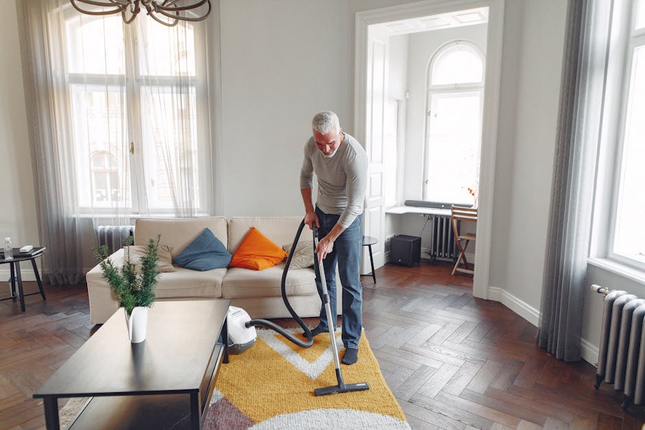 A man with gray hair and a beard is engaged in domestic cleaning, vacuuming a yellow and white patterned area rug in a bright living room with natural light streaming through large windows. The room features a white sofa adorned with colorful cushions (blue, orange, and beige), a dark coffee table with a potted plant, and wooden flooring arranged in a herringbone pattern. In the background, there is a small desk with a chair near a window, a radiator, and light-colored curtains. The scene emphasizes surface and deep cleaning, with a focus on maintaining hygiene and cleanliness, consistent with services offered by Carpet Cleaning Kennington, as indicated on the page titled 'Affordable Carpet Cleaning on Kennington Road and SE11, KENNINGTON'.