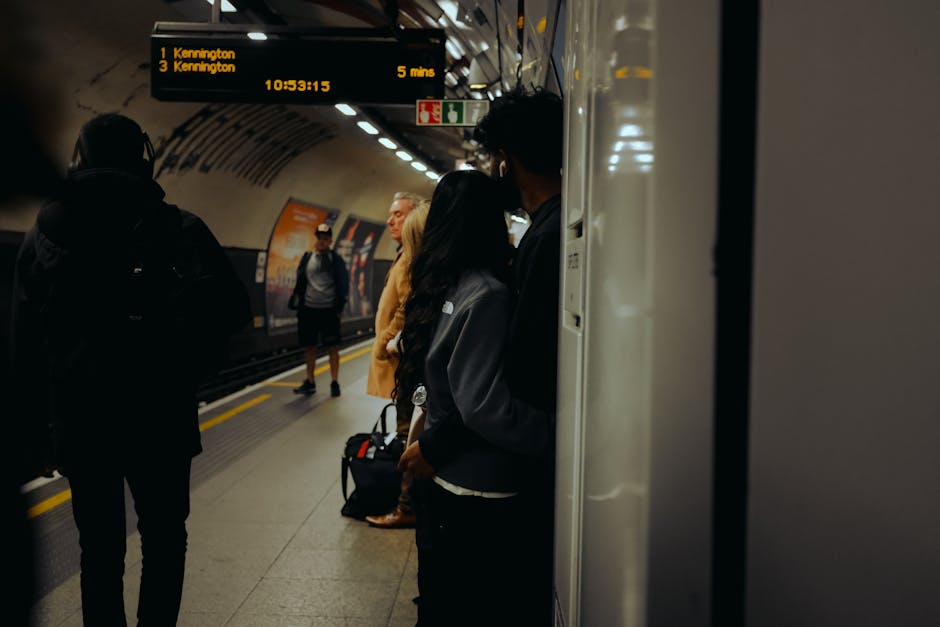 Image depicting a busy underground train station platform bathed in artificial lighting, with several commuters waiting for their train. The scene shows a mixture of individuals standing near the edge of the platform, some with backpacks and luggage. Visible surfaces include the tiled platform floor, which appears clean and well-maintained. A digital display overhead indicates the train times to Kennington and other destinations, with bright yellow text on a dark background. The station walls and ceiling are metallic with a curved design, and there are safety signs and advertisements posted along the platform. The overall scene emphasizes cleanliness and order typical of a professional cleaning environment, akin to the thorough surface cleaning and hygiene standards that Carpet Cleaning Kennington upholds in residential and commercial settings to avoid hidden charges in cleaning quotes. The lighting highlights the freshness of the space, suggesting regular sanitisation and deep cleaning practices that ensure a hygienic environment for passengers.