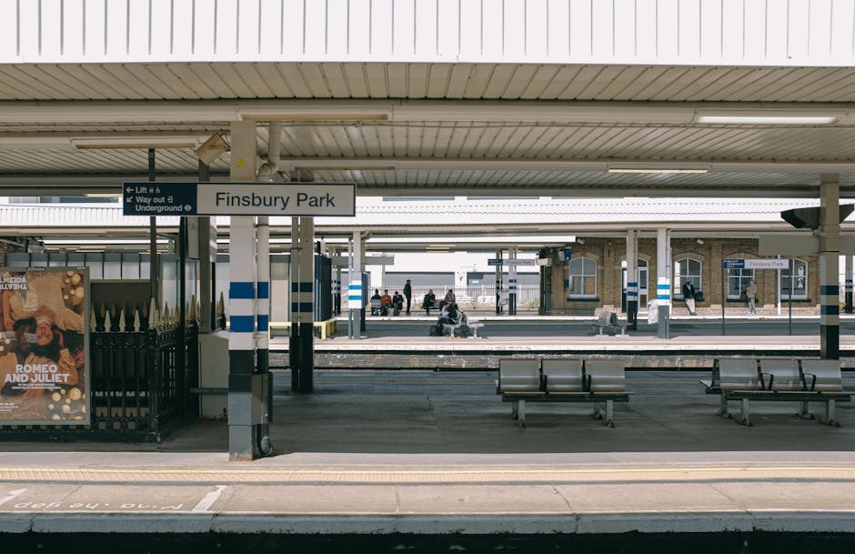 A view of Finsbury Park train station platform with a large white sign displaying the station name, 'Finsbury Park'. The platform surface appears clean and well-maintained, with metal benches for waiting passengers and a black metal fence on the left side. Overhead, a white corrugated roof covers the platform, providing shelter, with lighting fixtures embedded in the ceiling. In the background, multiple railway tracks extend through the station, and several passengers are seen sitting on benches, walking along the platform, or waiting for their trains. Small station buildings with brick facades and large windows are visible beyond the platform. The scene is illuminated by natural daylight, giving a clear and tidy impression of the station’s cleanliness, consistent with the high standards of surface cleaning and hygiene promoted by Carpet Cleaning Kennington, experts in domestic cleaning services near Kennington station.