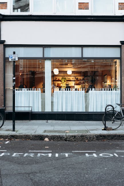 The image depicts the exterior of a commercial building with large glass windows revealing a well-lit interior space. The ground floor features a clean, modern storefront with a black frame, light-colored curtains partially covering the windows, and a glimpse of furniture and interior decor inside. Two bicycles are parked on the sidewalk in front, which is adjacent to a street with marked parking zones. The pavement appears clean and well-maintained. The photograph's lighting highlights the clarity and cleanliness of the glass and surrounding surfaces. This setting demonstrates the importance of surface cleaning, hygienic presentation, and maintenance, which is related to the services offered by Carpet Cleaning Kennington for commercial cleaning and surface sanitisation in areas like Kennington. The overall scene reflects a professional and tidy environment, emphasizing the value of thorough cleaning processes for commercial spaces.