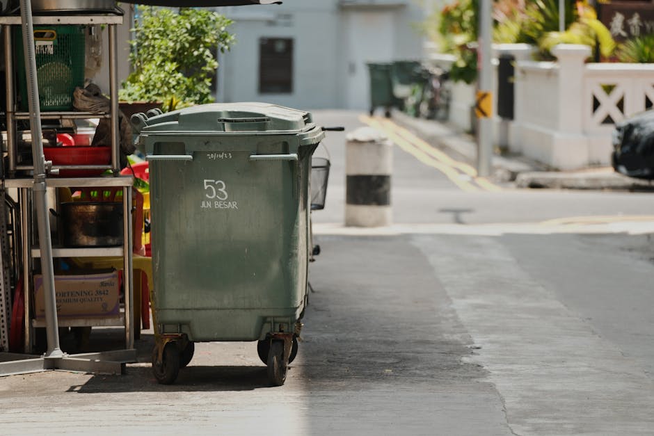 A green wheeled trash bin with white lettering marked '53 JUN BESAR' positioned on a sidewalk beside a storage rack filled with various cleaning tools and supplies, located near a curb on a residential street. The street features a concrete surface with minimal debris, with bollards and a section of road visible in the background. The area appears clean and orderly, with natural daylight illuminating the scene. The image depicts a typical waste disposal setup relevant to domestic or commercial cleaning practices, as referenced on the Lambeth Council Rules for Waste Disposal from Cleaning Jobs page, provided by Carpet Cleaning Kennington.