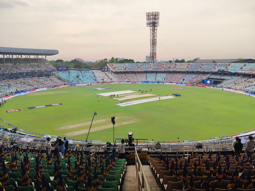 A view of a large cricket stadium during daytime, with the pitch covered by white tarpaulin sheets and surrounded by empty tiered seating areas. The seating includes rows of green and brown plastic chairs, some of which have colorful cloths draped over them. Bright floodlights are installed on tall towers above the stands, and a few individuals are present around the field, possibly event staff or media personnel. The stadium appears clean and well-maintained, with a clear view of the sky and a distant cityscape in the background. This setting illustrates a moment prior to or after a cricket match, with a focus on the arena's surface and seating area, indicative of professional sports event preparations, underlining the importance of surface cleaning and venue sanitation for safety and hygiene during sporting events, with Carpet Cleaning Kennington equipped to handle such maintenance tasks.