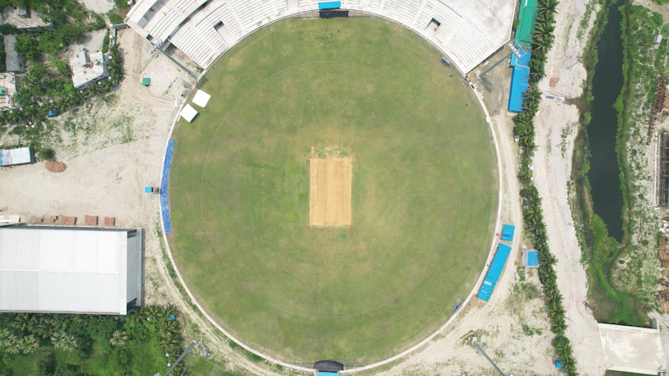A view of a large cricket stadium during daytime, with the pitch covered by white tarpaulin sheets and surrounded by empty tiered seating areas. The seating includes rows of green and brown plastic chairs, some of which have colorful cloths draped over them. Bright floodlights are installed on tall towers above the stands, and a few individuals are present around the field, possibly event staff or media personnel. The stadium appears clean and well-maintained, with a clear view of the sky and a distant cityscape in the background. This setting illustrates a moment prior to or after a cricket match, with a focus on the arena's surface and seating area, indicative of professional sports event preparations, underlining the importance of surface cleaning and venue sanitation for safety and hygiene during sporting events, with Carpet Cleaning Kennington equipped to handle such maintenance tasks.