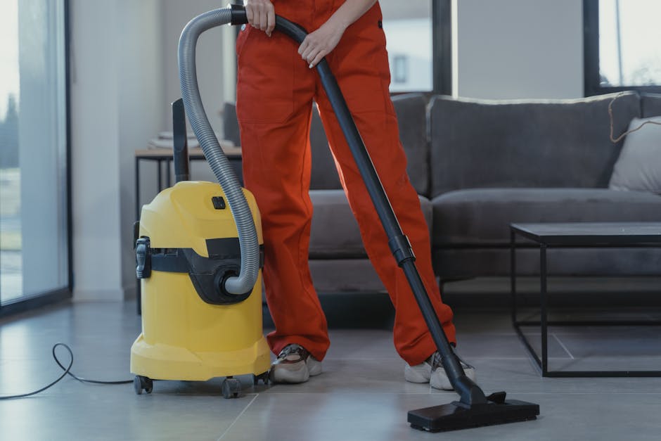 A person in orange overalls and white sneakers is operating a yellow commercial vacuum cleaner with a grey hose and black wand, performing surface cleaning on a tiled floor in a modern living room. Th