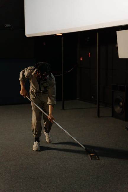 A person in orange overalls and white sneakers is operating a yellow commercial vacuum cleaner with a grey hose and black wand, performing surface cleaning on a tiled floor in a modern living room. Th