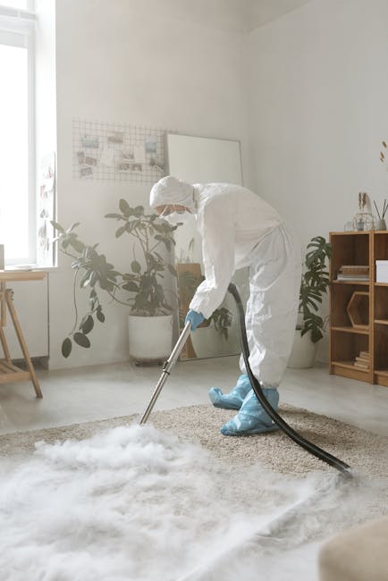 A professional cleaner from Carpet Cleaning Kennington is performing a deep surface cleaning on a light beige or cream-colored carpet in a well-lit, modern living room. The cleaner is wearing full protective gear, including a white suit, gloves, face mask, and blue shoe covers, and is using a black vacuum or extraction tool connected to a hose to ensure thorough sanitisation of the carpet, which appears damp during the cleaning process. The room features white walls, a large window allowing natural light, a wooden sideboard with minimal decorative items, and several green houseplants, creating a clean and tidy atmosphere focused on hygiene and maintenance. The scene exemplifies domestic cleaning and surface deep cleaning aimed at removing stains, dust, and dirt to ensure a hygienic environment, consistent with the services offered by Carpet Cleaning Kennington for emergency stain removal and quick turnaround cleaning needs.