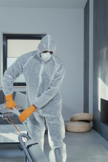 A cleaning professional wearing a white protective coverall, face mask, and orange gloves uses a portable vacuum or cleaning device to disinfect a modern residential room with large windows, grey walls, and a tiled floor. The room is well-lit with natural light, and there are two rounded woven cushions on the floor near the window. The scene emphasizes thorough surface cleaning, sanitisation, and maintenance, showcasing modern domestic cleaning practices as provided by Carpet Cleaning Kennington for effective hygiene and deep cleaning in a home environment.