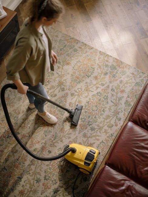 A professional cleaner from Carpet Cleaning Kennington is performing a deep surface cleaning on a light beige or cream-colored carpet in a well-lit, modern living room. The cleaner is wearing full protective gear, including a white suit, gloves, face mask, and blue shoe covers, and is using a black vacuum or extraction tool connected to a hose to ensure thorough sanitisation of the carpet, which appears damp during the cleaning process. The room features white walls, a large window allowing natural light, a wooden sideboard with minimal decorative items, and several green houseplants, creating a clean and tidy atmosphere focused on hygiene and maintenance. The scene exemplifies domestic cleaning and surface deep cleaning aimed at removing stains, dust, and dirt to ensure a hygienic environment, consistent with the services offered by Carpet Cleaning Kennington for emergency stain removal and quick turnaround cleaning needs.