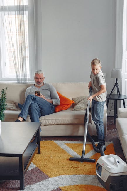 A living room scene featuring a light beige sofa with orange and neutral cushions, situated against a light grey wall with large windows draped in sheer curtains allowing natural daylight to illuminate the space. A man with grey hair and beard, dressed in a grey long-sleeve shirt and blue jeans, is seated on the sofa, looking at a smartphone. A young boy with blond hair, wearing a grey t-shirt and jeans, is standing nearby operating a vacuum cleaner on a multicolored rug with geometric patterns in yellow, white, orange, and purple. A black side table holding a white lamp is positioned beside the sofa. The room appears clean and well-maintained, with the vacuum cleaner in use indicating ongoing surface cleaning or deep cleaning activity, which aligns with expert domestic cleaning services provided by Carpet Cleaning Kennington.