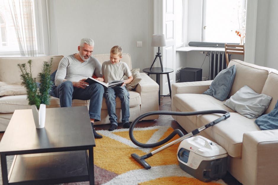 A spacious living room with natural daylight coming through large white-framed windows, showcasing a beige fabric sofa with blue and grey cushions, a black round side table with a lamp, and a wooden chair positioned near the window. In the foreground, a modern vacuum cleaner with a black hose and white body rests on a multicolored geometric-patterned area rug, indicating recent cleaning activity. The room features light grey walls, wooden flooring, and minimalistic decor, highlighting the clean and well-maintained condition of the space. Carpet Cleaning Kennington's services are reflected in the emphasis on surface cleaning and deep sanitisation for residential environments.