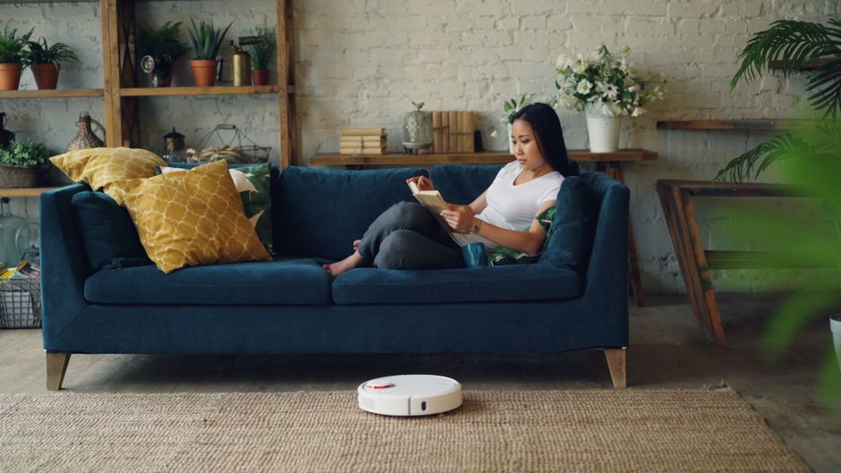 A living room scene featuring a light beige sofa with orange and neutral cushions, situated against a light grey wall with large windows draped in sheer curtains allowing natural daylight to illuminate the space. A man with grey hair and beard, dressed in a grey long-sleeve shirt and blue jeans, is seated on the sofa, looking at a smartphone. A young boy with blond hair, wearing a grey t-shirt and jeans, is standing nearby operating a vacuum cleaner on a multicolored rug with geometric patterns in yellow, white, orange, and purple. A black side table holding a white lamp is positioned beside the sofa. The room appears clean and well-maintained, with the vacuum cleaner in use indicating ongoing surface cleaning or deep cleaning activity, which aligns with expert domestic cleaning services provided by Carpet Cleaning Kennington.